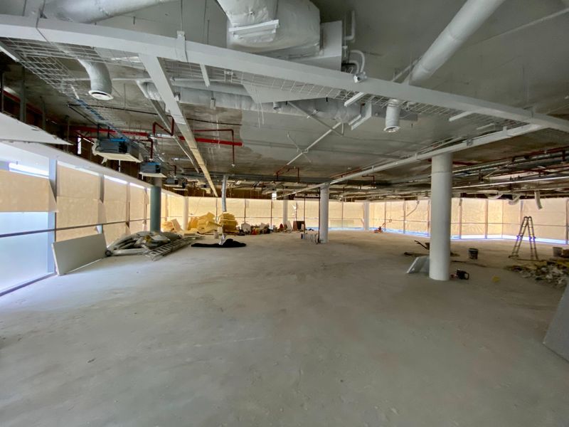 A large, open-plan commercial office space currently under construction, featuring exposed ceilings, structural columns, and expansive floor-to-ceiling windows.