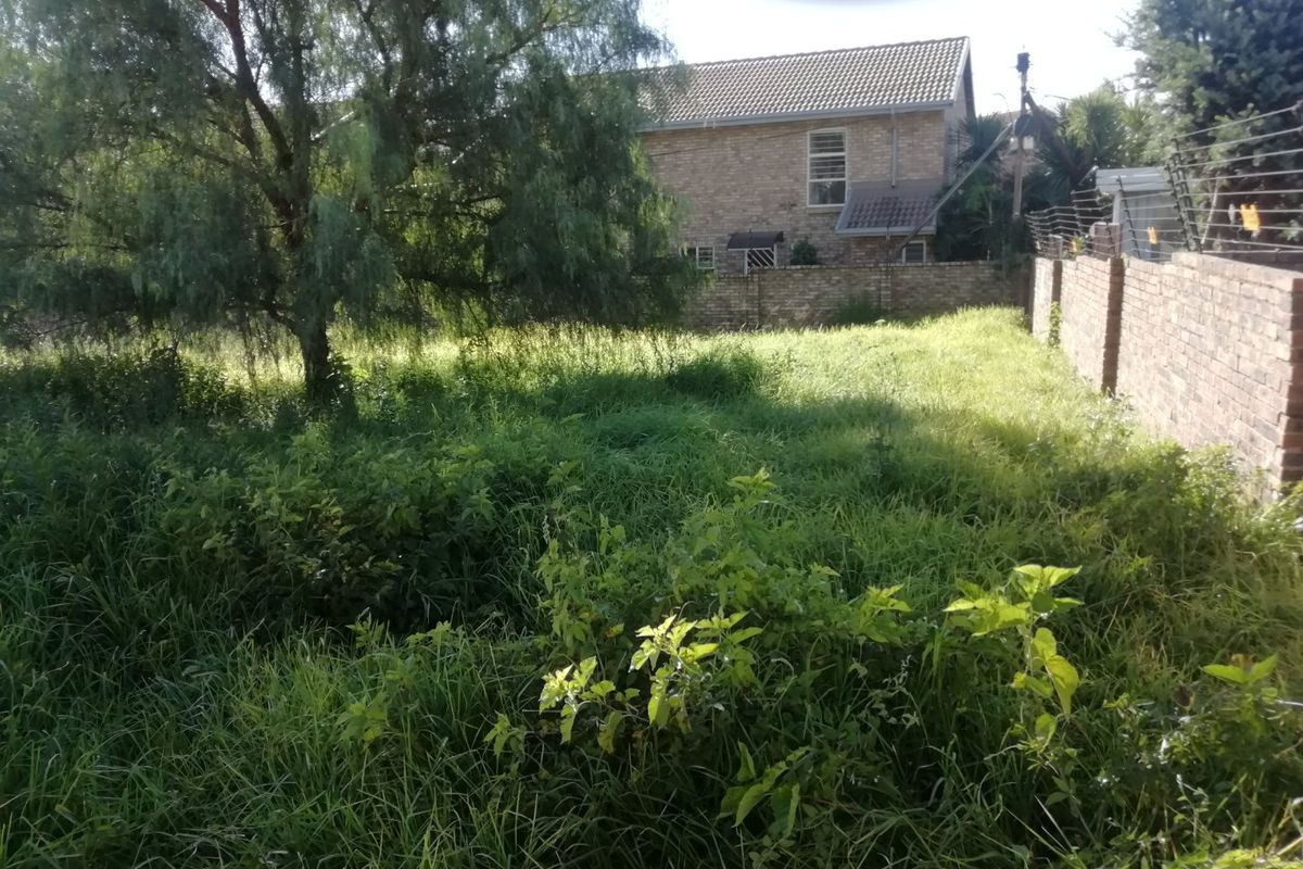 A view of a vacant residential plot featuring overgrown grass, mature trees, and a brick boundary wall with electric fencing.