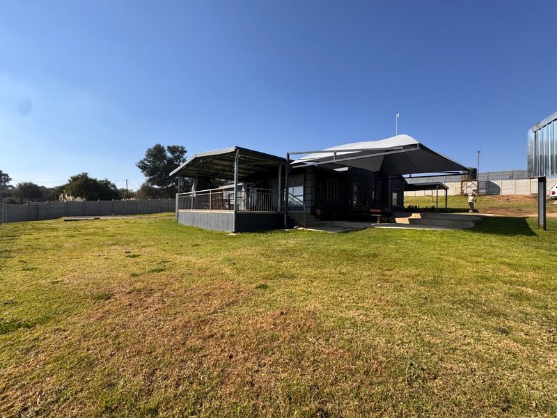 Rear view of the property featuring a large lawn, covered patio area with metal roofing, and perimeter concrete walling.