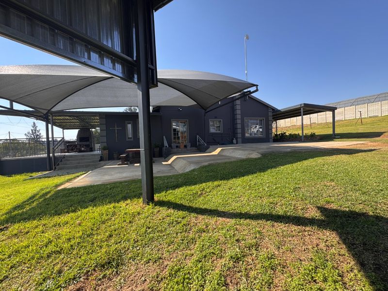 Rear exterior view featuring a spacious concrete patio, shade sail covering, and a built-in braai area set against a well-maintained lawn.