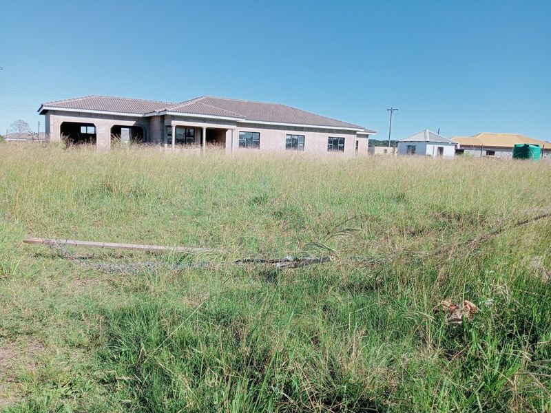 A view of an unfinished residential property featuring a tiled roof and plastered exterior, situated on a large, overgrown plot of land.