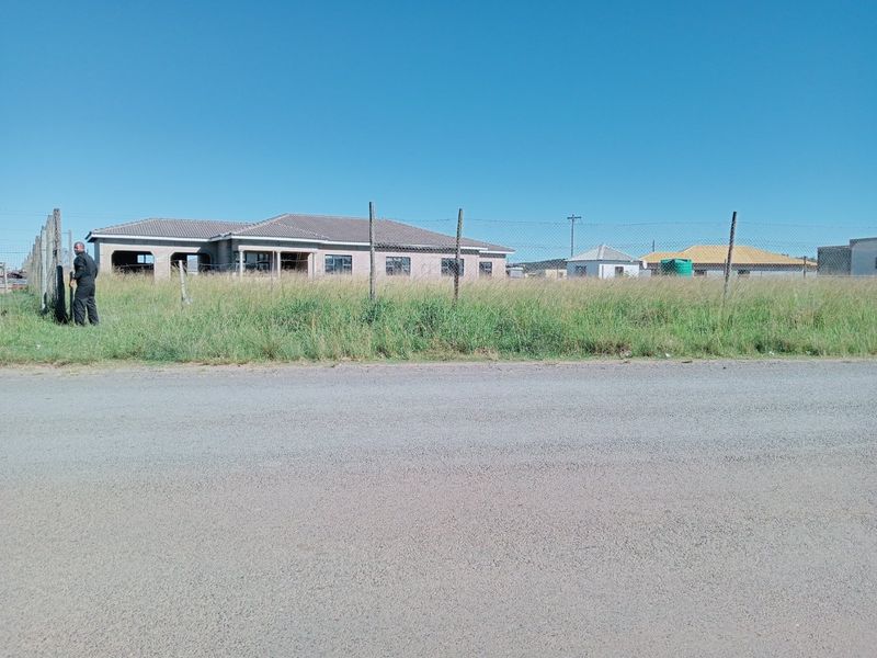 Street view of a residential property under construction, featuring a tiled roof and wire fencing with overgrown grass in the foreground.