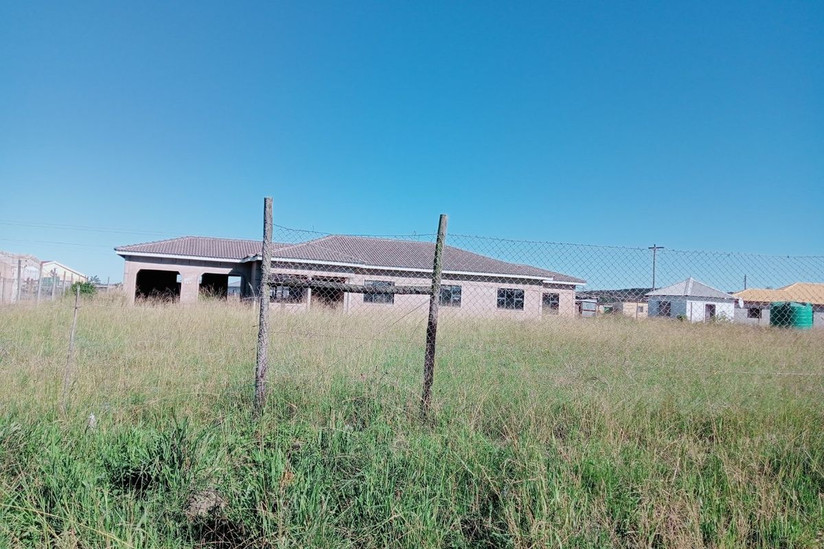 An exterior view of an unfinished residential property featuring a tiled roof and concrete block structure, set on a large plot with perimeter fencing.