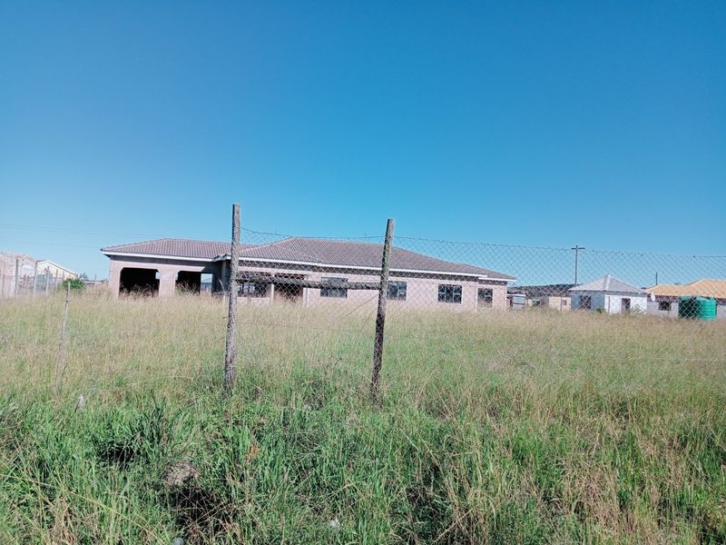 An exterior view of an unfinished residential property featuring a tiled roof and concrete block structure, set on a large plot with perimeter fencing.