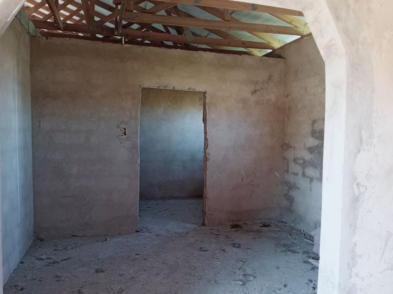 An unfinished interior room featuring exposed timber roof trusses, plastered masonry walls, and an arched doorway, currently in a shell construction state.