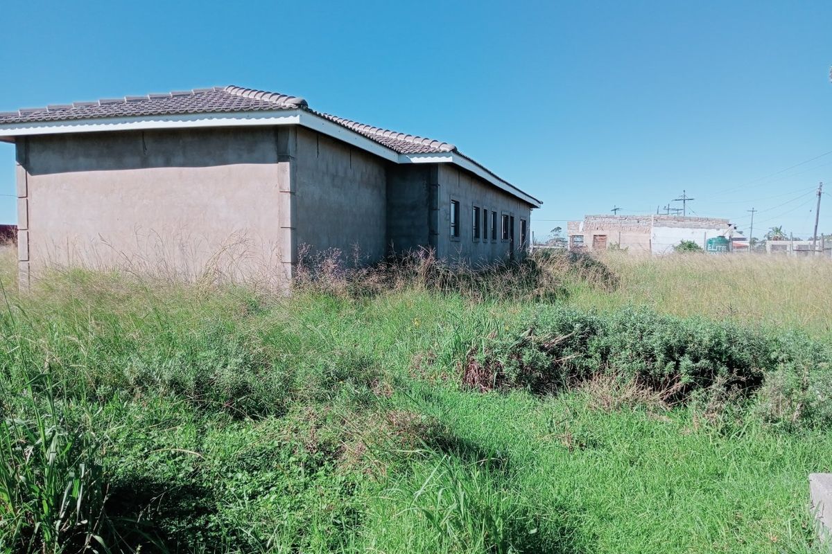 Exterior view of a single-story house with a tiled roof and plastered walls, surrounded by an overgrown garden area.