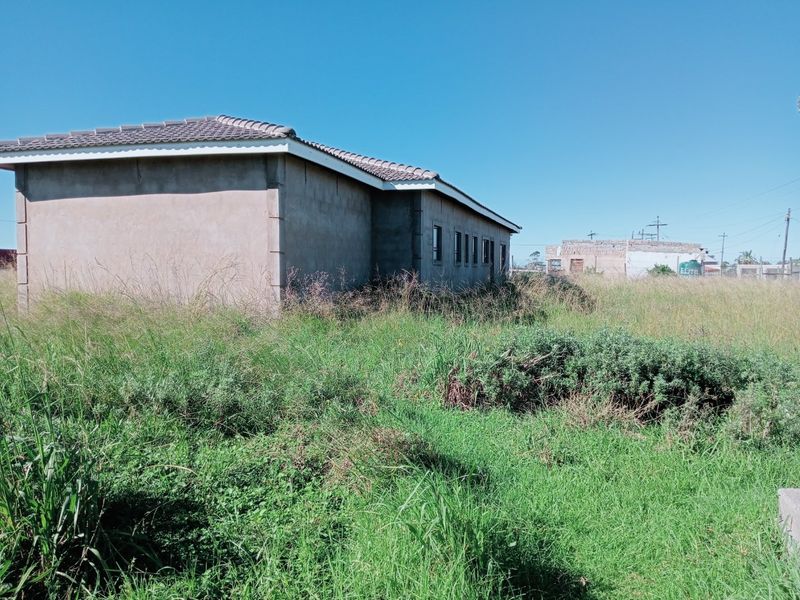 Exterior view of a single-story house with a tiled roof and plastered walls, surrounded by an overgrown garden area.