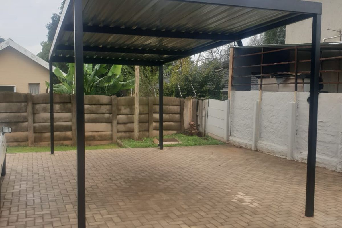 A well-maintained metal carport structure situated over a paved driveway, enclosed by precast concrete boundary walls in a rear yard setting.