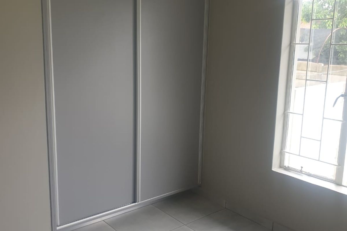 A clean, empty bedroom featuring neutral tiled flooring, a modern built-in sliding door wardrobe, and a window fitted with security burglar bars.