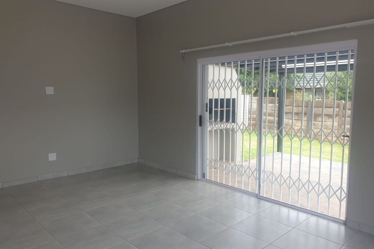 A bright, empty living area featuring neutral tiled flooring and a sliding glass door with a security gate leading to the outdoor patio.