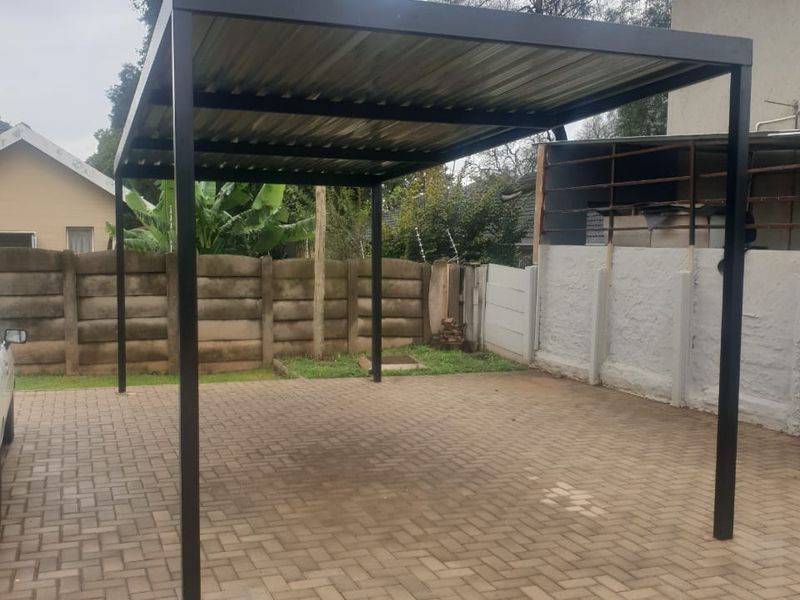 A well-maintained metal carport structure situated over a paved driveway, enclosed by precast concrete boundary walls in a rear yard setting.