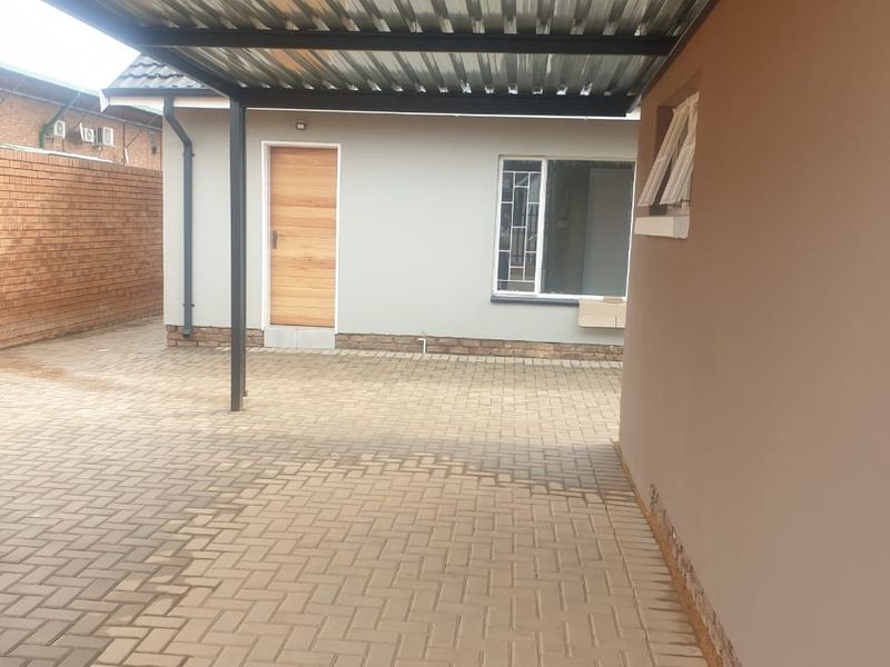 A paved rear courtyard area featuring a covered carport structure, brick boundary wall, and access to the main building via a timber door.