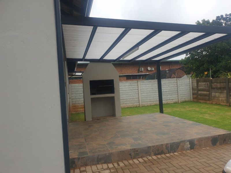 A covered outdoor patio area featuring a built-in braai, tiled flooring, and a modern pergola structure, overlooking a private garden space.
