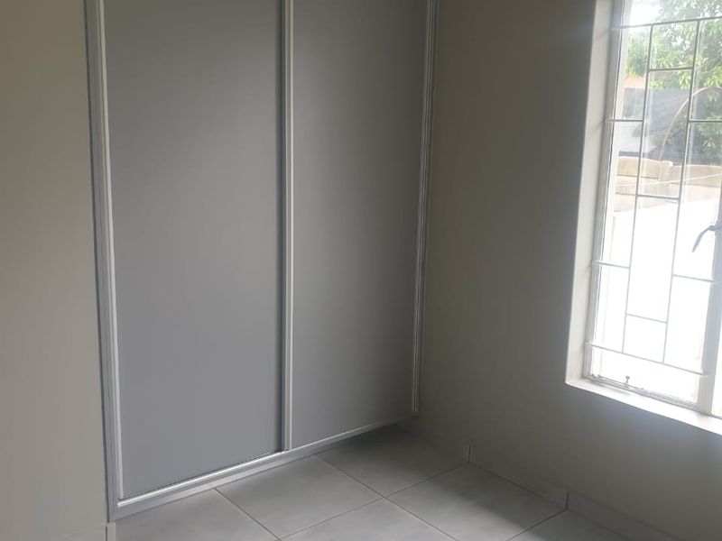 A clean, empty bedroom featuring neutral tiled flooring, a modern built-in sliding door wardrobe, and a window fitted with security burglar bars.