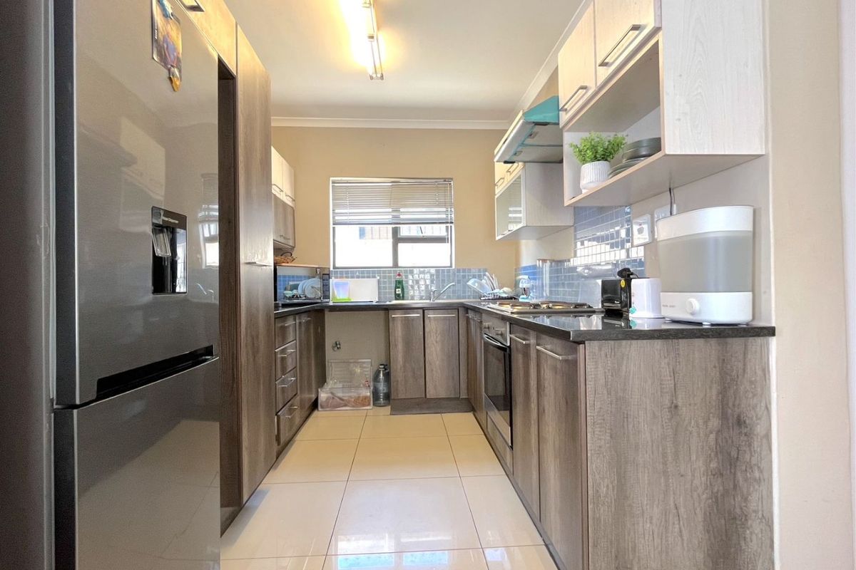 A functional galley-style kitchen featuring wood-look cabinetry, stone countertops, a tiled backsplash, and integrated cooking appliances with neutral floor tiles.