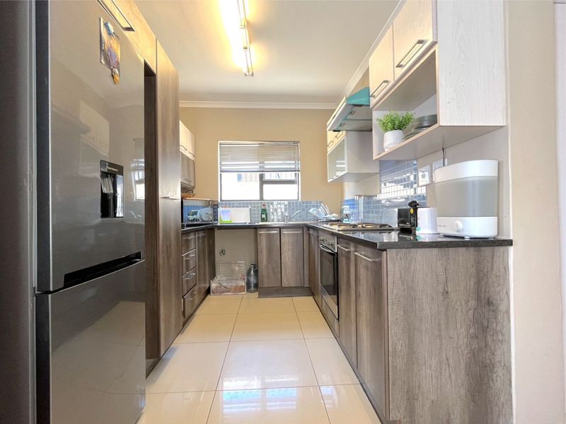 A functional galley-style kitchen featuring wood-look cabinetry, stone countertops, a tiled backsplash, and integrated cooking appliances with neutral floor tiles.