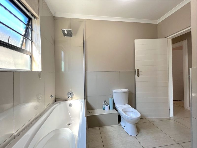 A clean, modern bathroom featuring a built-in bathtub with an overhead shower, a toilet, and neutral-toned wall and floor tiles.