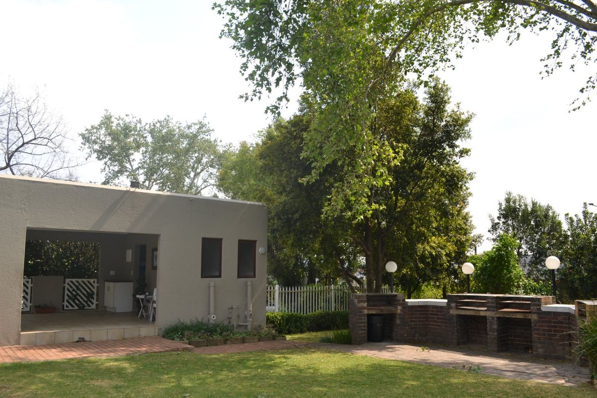 Rear garden area featuring a covered patio, built-in brick braai, paved pathways, and established trees within a fenced yard.