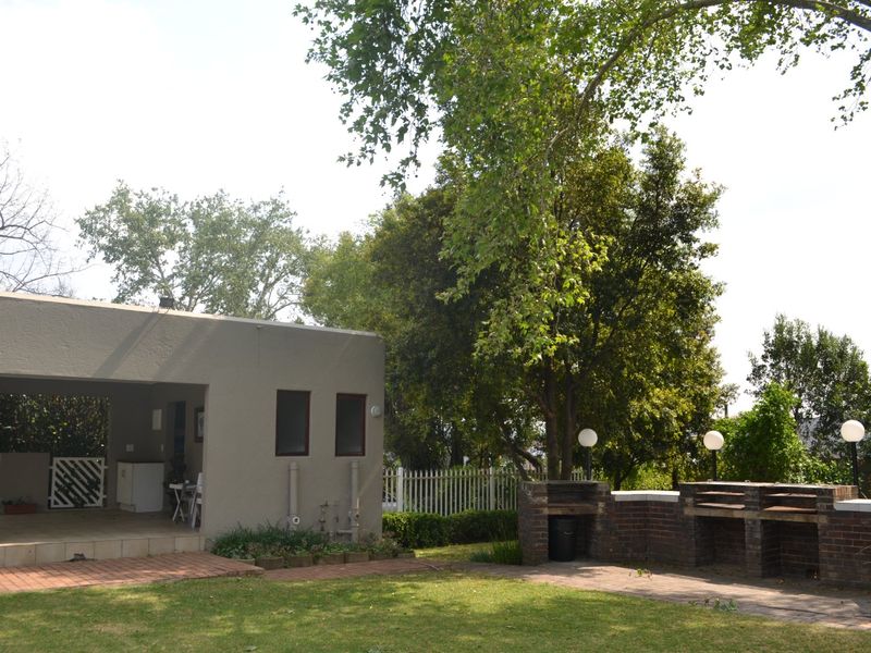Rear garden area featuring a covered patio, built-in brick braai, paved pathways, and established trees within a fenced yard.