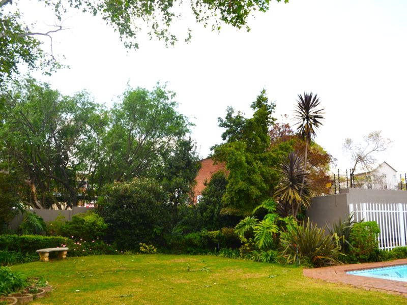 A well-maintained garden area featuring a swimming pool with brick coping, surrounded by established trees, shrubs, and a secure perimeter wall with electric fencing.