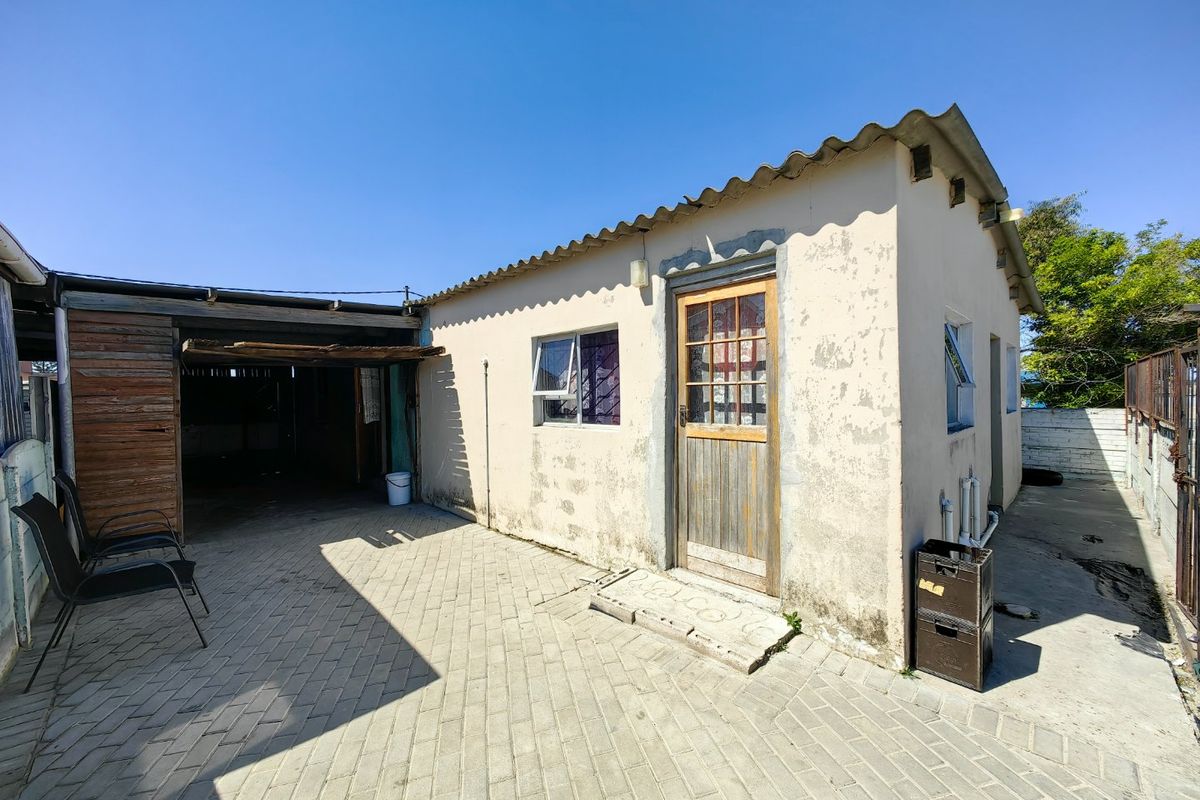 Rear exterior view of the property featuring a paved courtyard, covered carport area, and a building with weathered exterior finishes.