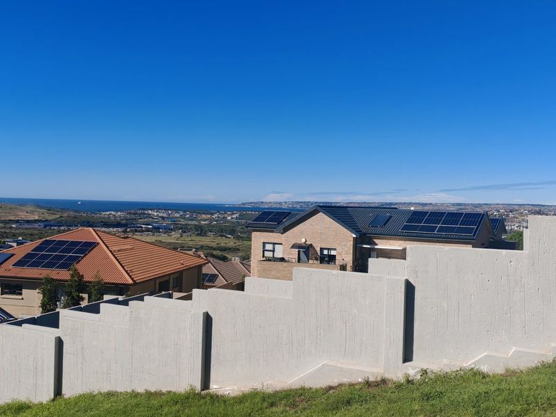 Elevated view overlooking the residential neighborhood and coastline, featuring prominent concrete retaining walls and houses equipped with solar panel installations.