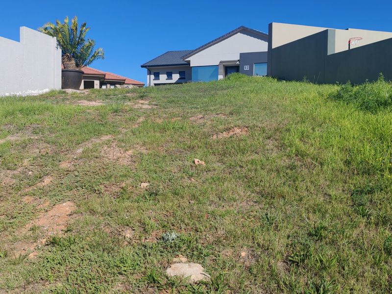 A view of a vacant residential plot featuring a sloped grassy terrain and surrounding boundary walls in a developed neighborhood.