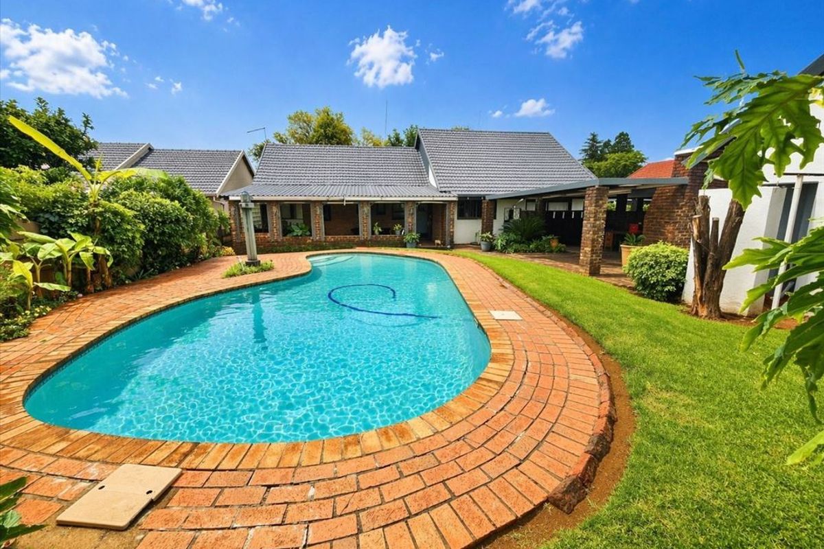 A well-maintained backyard featuring a swimming pool with brick coping, a covered patio area, and established garden landscaping.