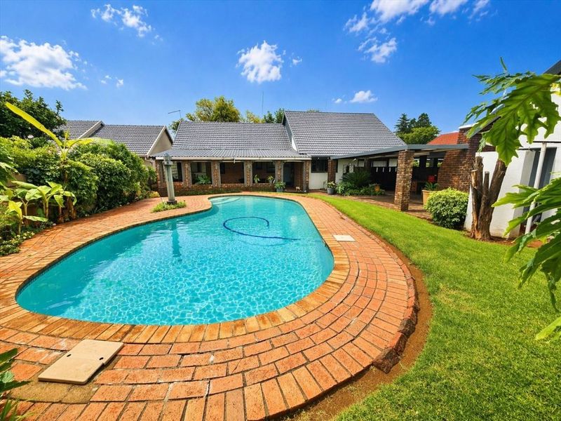 A well-maintained backyard featuring a swimming pool with brick coping, a covered patio area, and established garden landscaping.