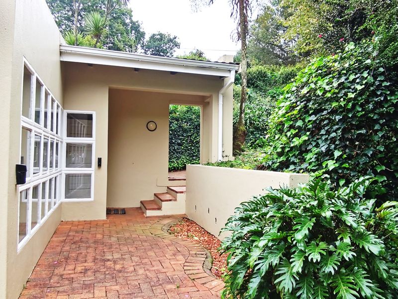 A private, paved outdoor patio area featuring a covered walkway, brick flooring, and established garden surroundings.