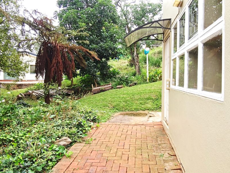 A rear exterior view featuring a brick paved pathway leading along the side of the house, bordered by a garden area with mature trees.