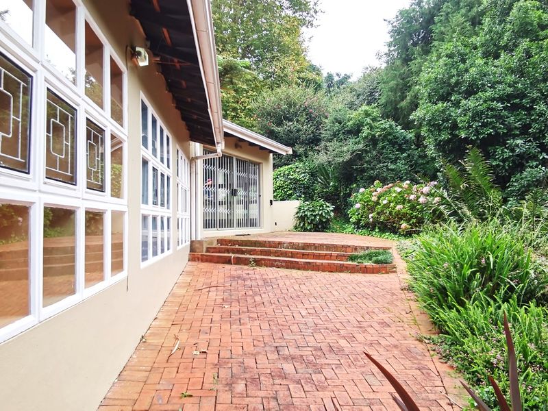 Rear exterior view featuring a brick paved patio, raised brick steps, and large multi-pane windows leading into the home, surrounded by established garden greenery.