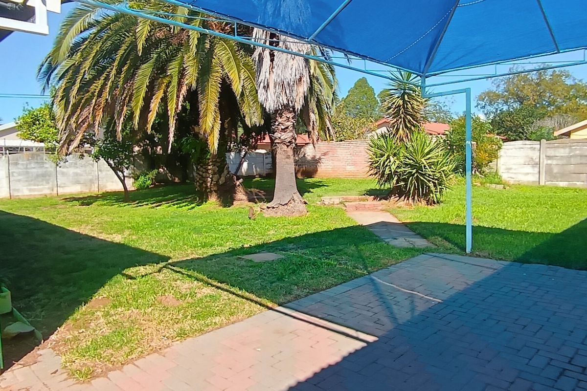 A spacious backyard featuring a paved patio area with a blue shade sail, mature palm trees, and a well-maintained lawn enclosed by boundary walls.