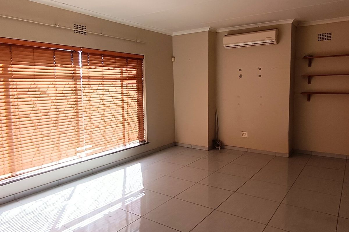A spacious, empty living room featuring neutral tiled flooring, wooden venetian blinds, wall-mounted air conditioning unit, and built-in wooden shelving.