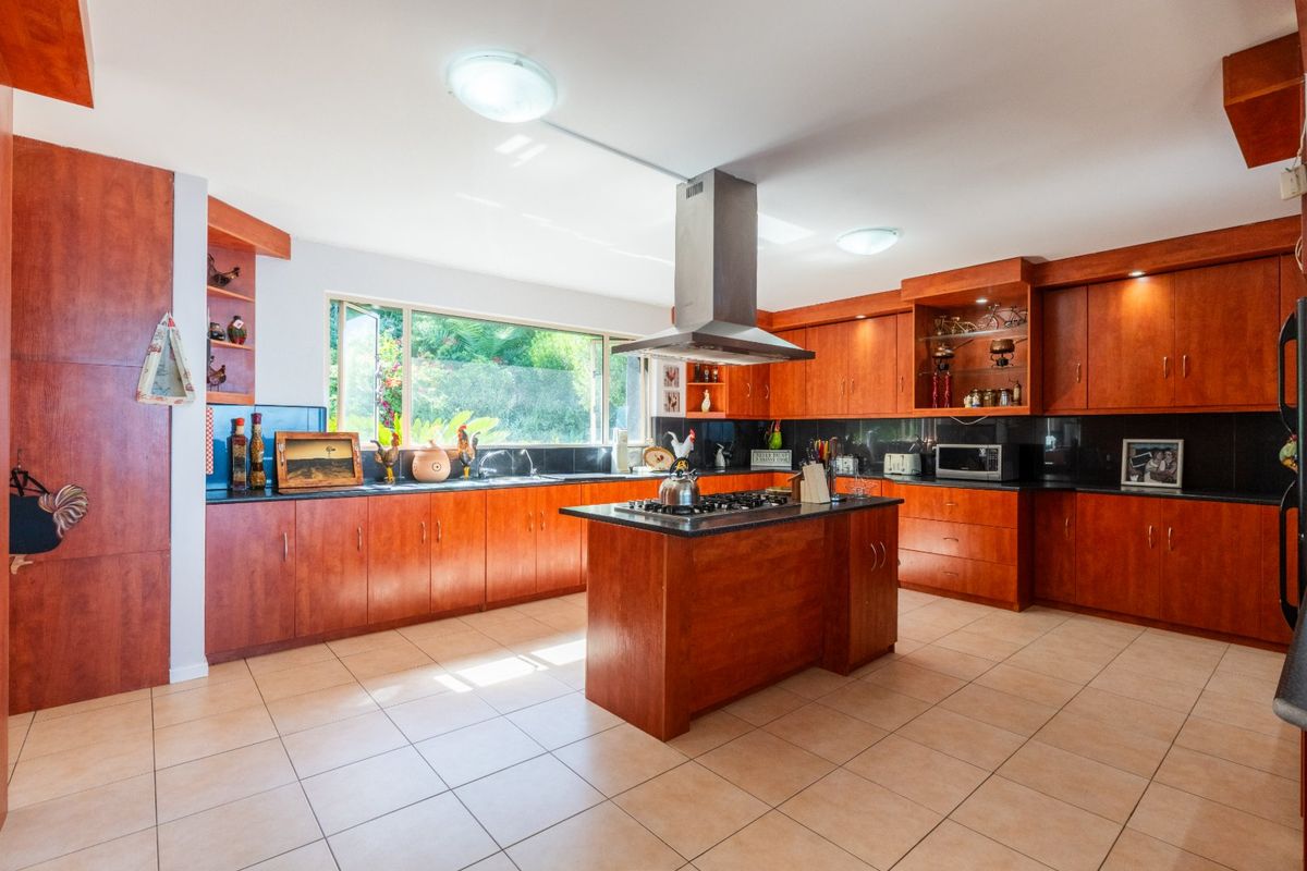 Spacious kitchen featuring warm-toned wooden cabinetry, a central island with a gas cooktop, stainless steel extractor fan, and large windows overlooking the garden.