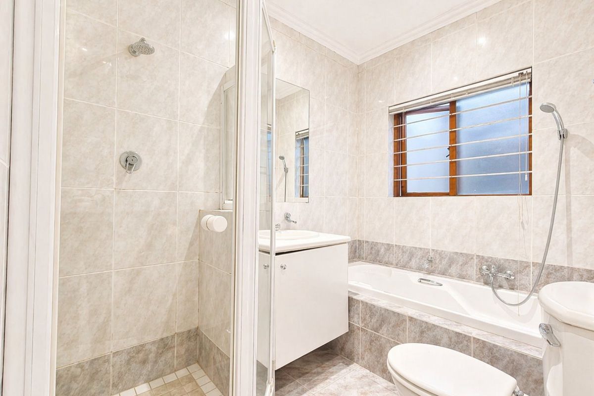 A functional bathroom featuring a glass-enclosed shower, a built-in bathtub, a vanity basin, and a toilet, finished with neutral wall and floor tiling.