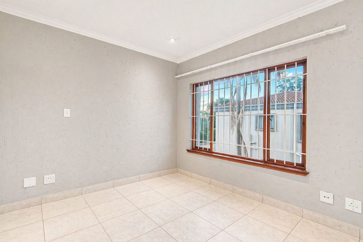 A bright, empty bedroom featuring neutral tiled flooring, textured grey walls, recessed ceiling lighting, and a large window with security bars.