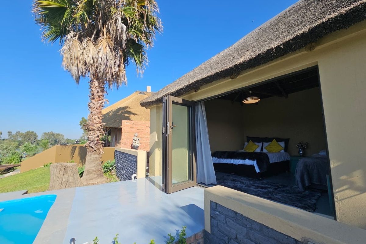 A bedroom with bi-fold doors opening onto a patio area with a swimming pool, featuring a traditional thatch roof design.