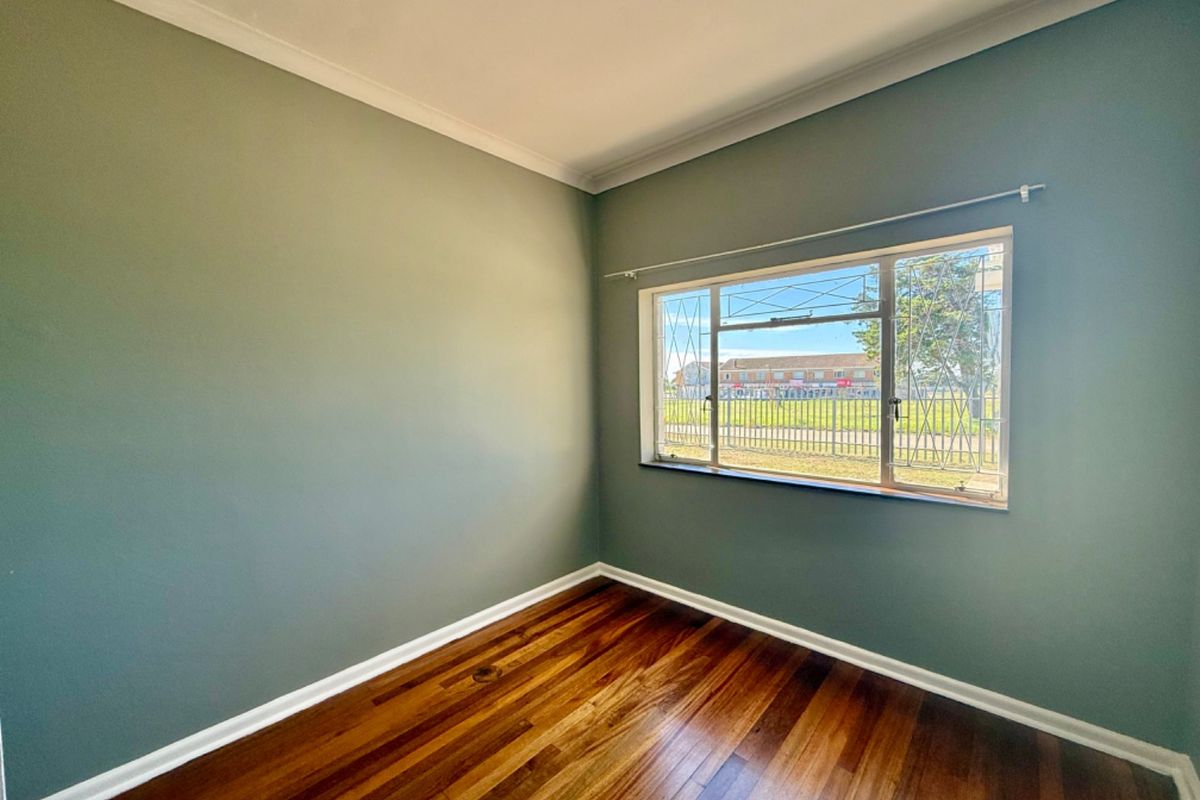 A bright, empty bedroom featuring polished hardwood flooring, neutral green walls, and a window fitted with security bars.
