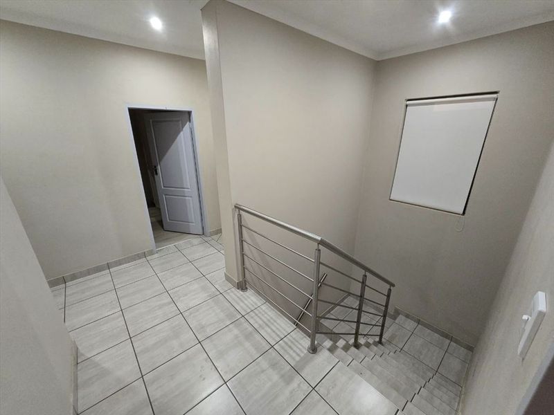 A clean, modern hallway featuring tiled flooring, recessed downlighting, and a stainless steel staircase balustrade leading to the lower level.