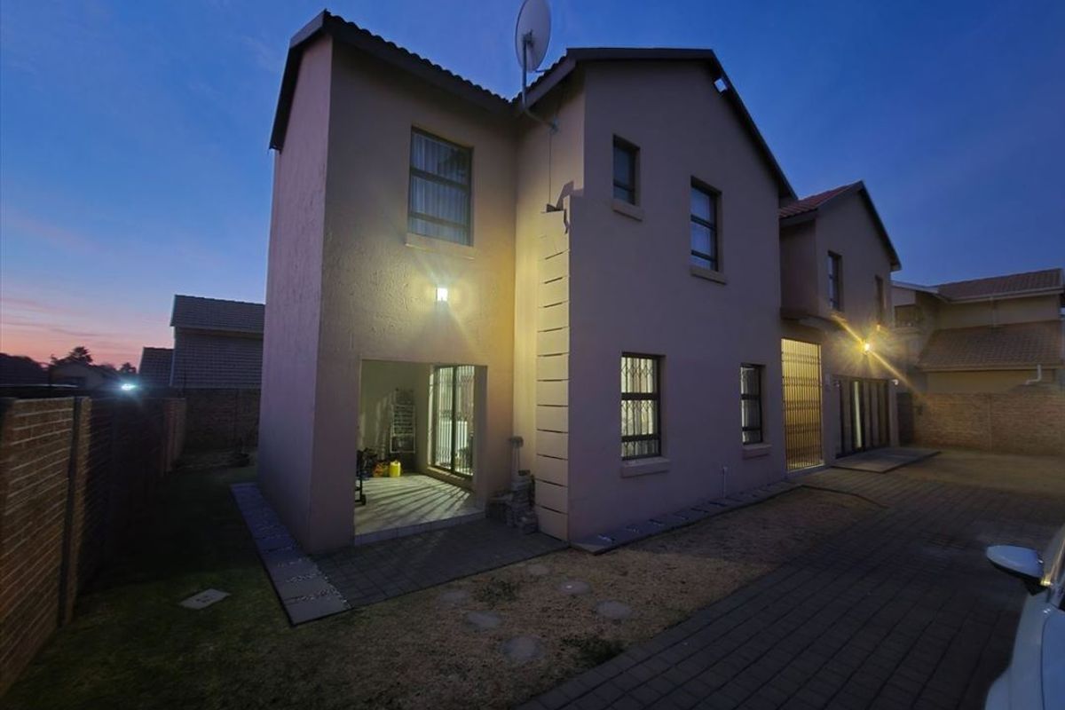 Rear view of the double-story residential property featuring paved driveway, security gates, and exterior wall lighting at dusk.