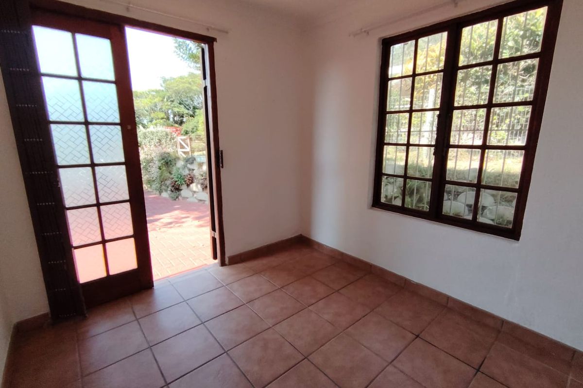 A bright, empty living space featuring terracotta floor tiles, timber-framed windows, and a glass-paneled door opening onto an outdoor patio area.