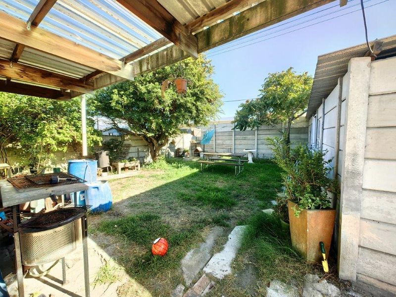 A covered outdoor patio area featuring a corrugated roof structure and timber beams, overlooking a private garden with precast concrete boundary walls.