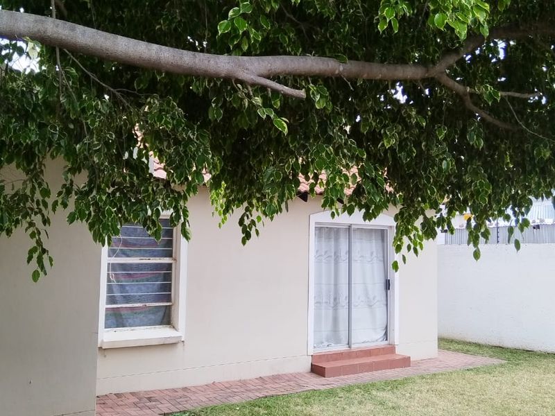 A view of the rear exterior of the house featuring a paved walkway, sliding glass doors, and a large mature tree providing shade.