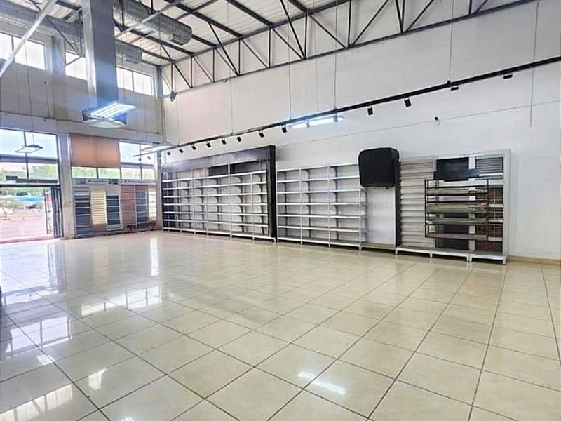 Spacious commercial open-plan showroom featuring high ceilings with exposed industrial ducting, tiled flooring, built-in retail shelving, and large glass windows for natural light.