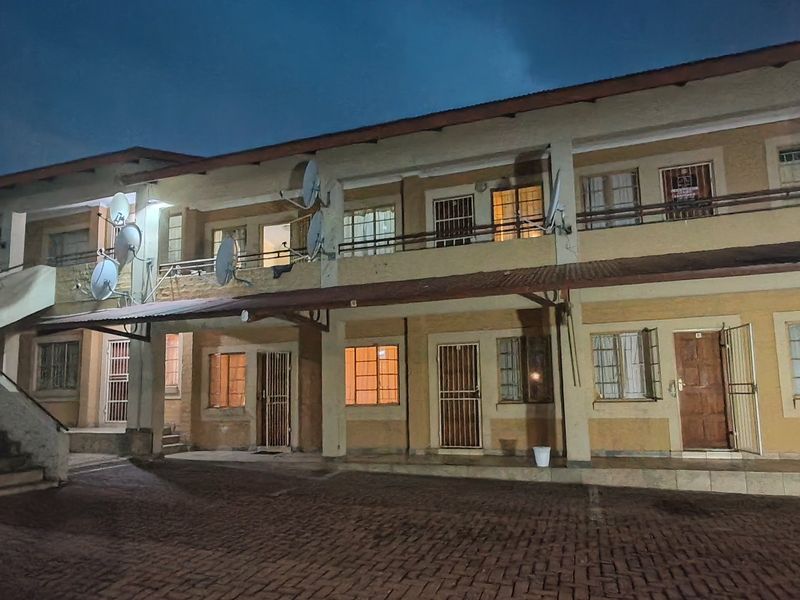 Exterior view of a multi-story apartment complex at dusk, featuring paved driveway access, security gates, and balcony access for upper-level units.