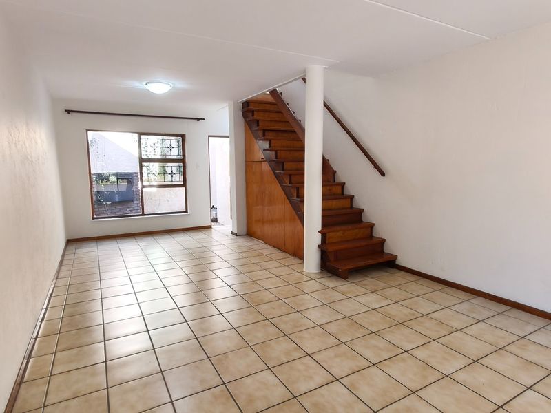 An empty, open-plan living area featuring tiled flooring, a wooden staircase with integrated under-stair storage, and a large window with security bars.