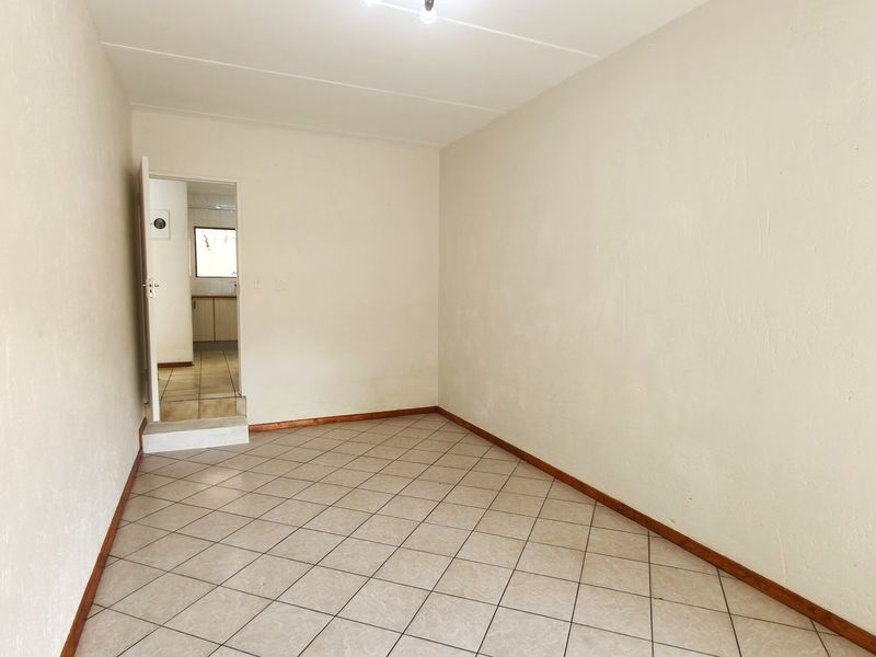 An empty, neutrally painted living area featuring tiled flooring and skirting boards, with a doorway leading to an adjacent kitchen space.