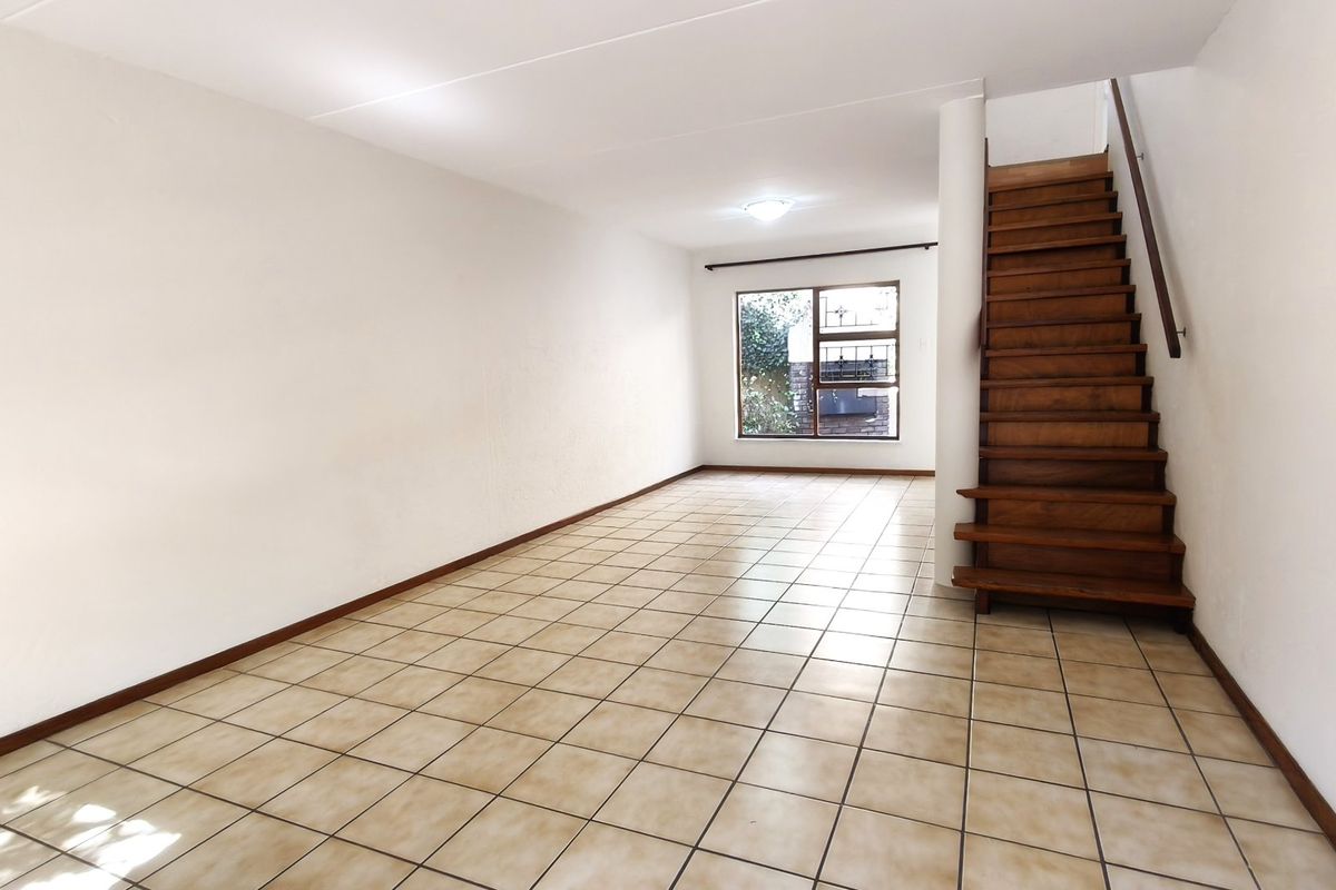 An empty, open-plan living area featuring tiled flooring and a wooden staircase leading to the upper level, with natural light from a rear window.