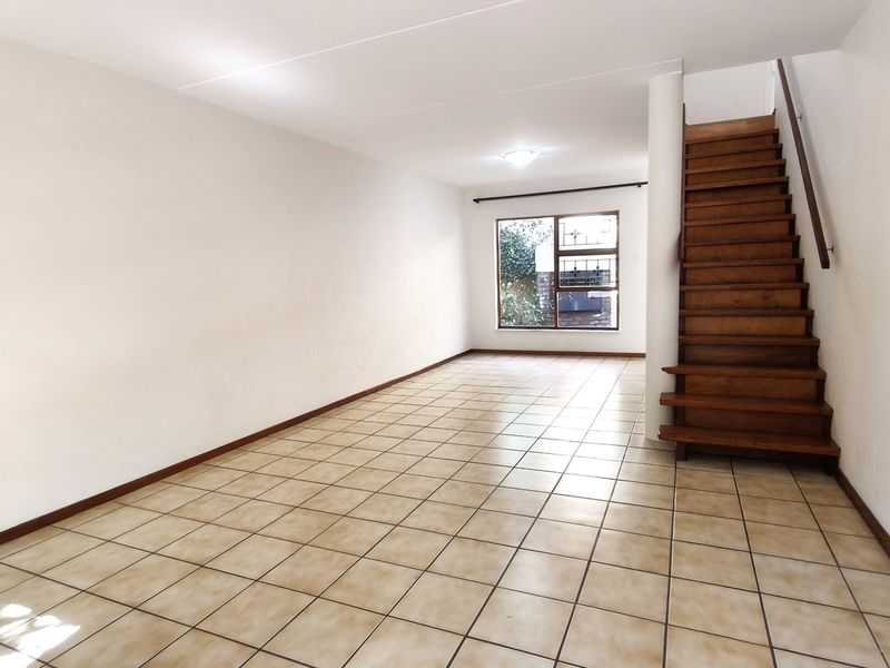 An empty, open-plan living area featuring tiled flooring and a wooden staircase leading to the upper level, with natural light from a rear window.